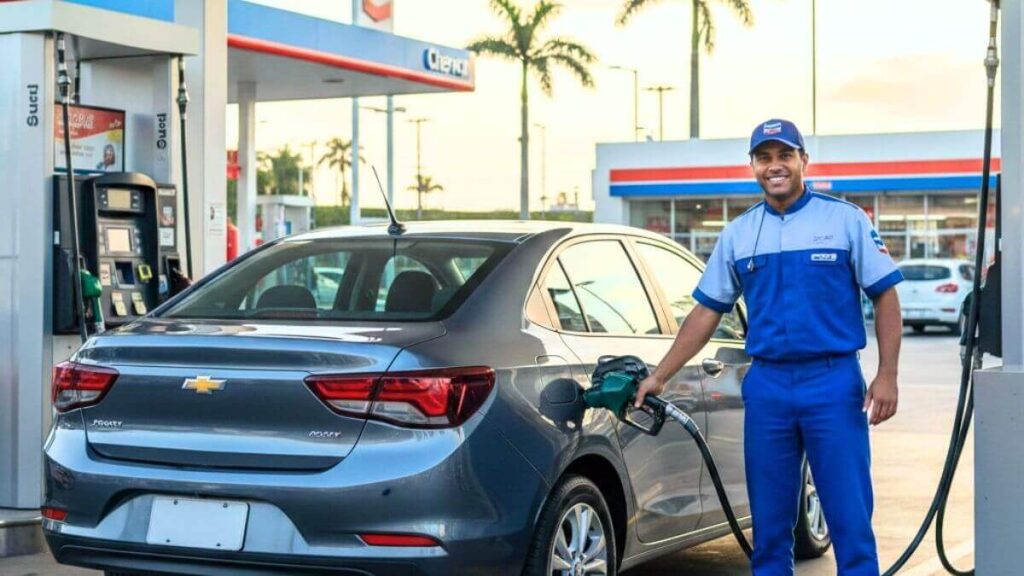 Um Chevrolet Onix de cor cinza estacionado em uma bomba de combustível, enquanto um frentista uniformizado e sorridente segura a mangueira de abastecimento.