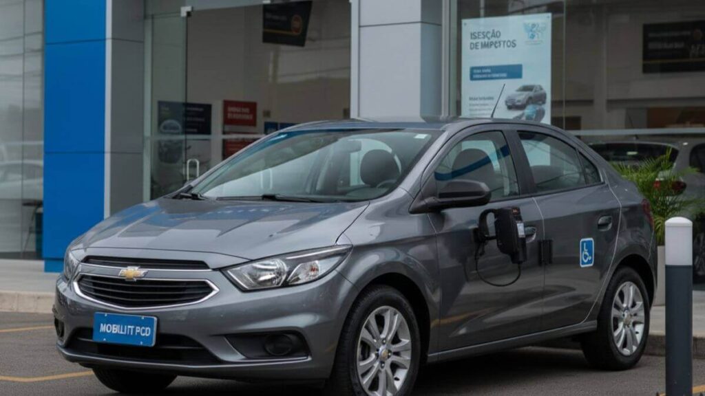 Chevrolet Onix cinza com adesivo de acessibilidade (PCD) estacionado em frente a uma concessionária moderna com vitrines de vidro, destacando o direito à isenção de impostos.