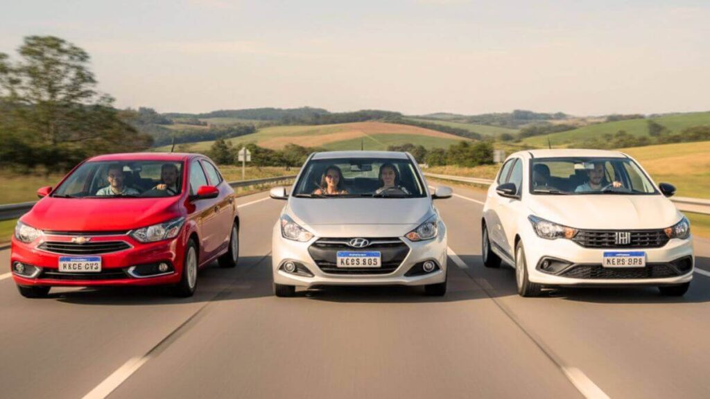 Três carros em movimento em uma estrada asfaltada: um Chevrolet Onix vermelho, um Hyundai HB20 prata e um Fiat Argo branco. Os motoristas (homem e mulher) aparecem sorrindo através do para-brisa, transmitindo sensação de conforto e prazer ao dirigir.