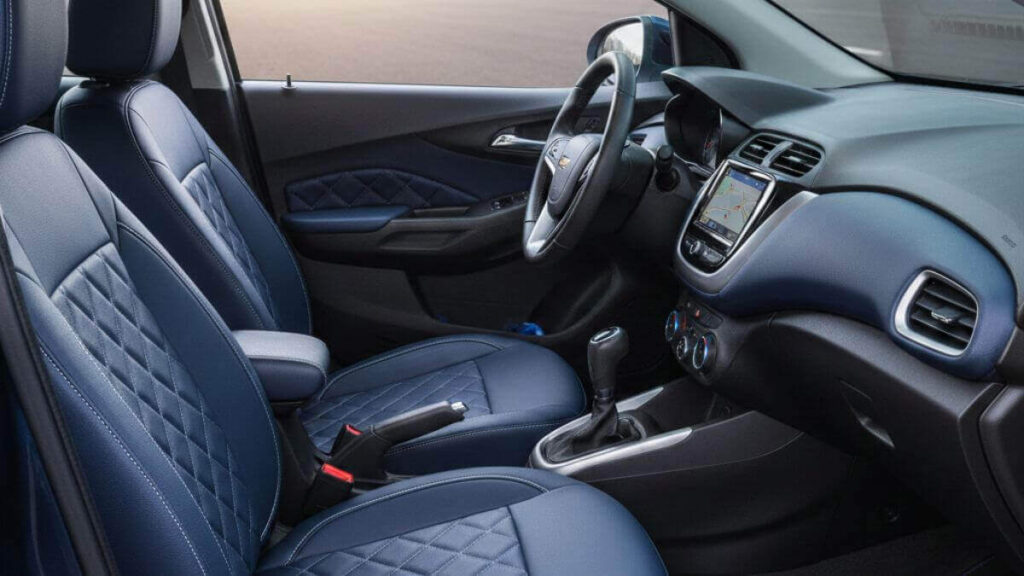 Visão interna de um Chevrolet Onix azul com foco nos bancos dianteiros revestidos em couro sintético automotivo azul marinho e acabamento em costura diamante (matelassê). O painel e o console central são visíveis em um ambiente iluminado e profissional.