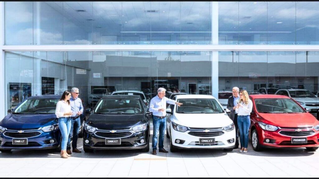 Fileira de carros Chevrolet Onix nas cores azul, preto, branco e vermelho estacionados em frente a uma concessionária moderna, com pessoas ao redor admirando os veículos.