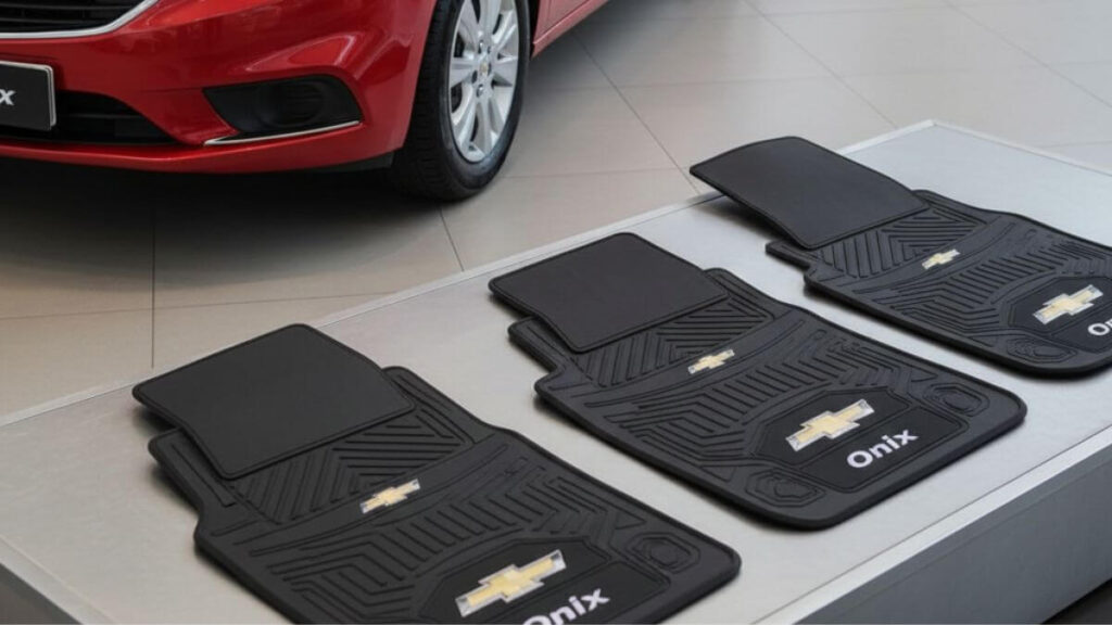 Foto publicitária de um Chevrolet Onix vermelho brilhante estacionado, com um conjunto de quatro tapetes automotivos personalizados exibidos ao lado, sobre um suporte elegante. Os tapetes possuem bordado com o logotipo da Chevrolet e o nome Onix.