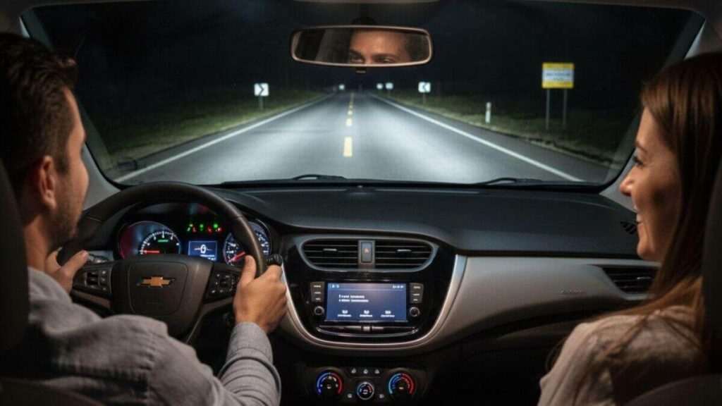 Vista de dentro de um Chevrolet Onix à noite, mostrando o painel moderno e a estrada à frente fortemente iluminada pelos faróis do veículo.