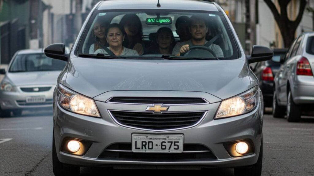 Vista frontal de um Chevrolet Onix prata 2015 subindo uma rua inclinada em um ambiente urbano. O veículo está ocupado por motorista e passageiros. No espelho retrovisor interno, destaca-se um aviso luminoso verde indicando 'A/C Ligado', simbolizando o esforço mecânico extra do motor em subidas.