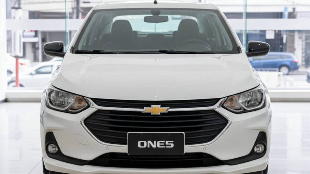 Visão frontal de um Chevrolet Onix 2026 na cor branca, versão aspirada, posicionado dentro de uma concessionária iluminada, mostrando a grade frontal, faróis e o emblema da Chevrolet.