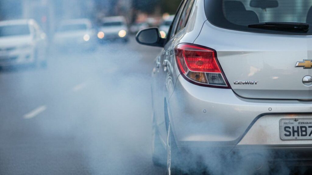 Vista traseira de um Chevrolet Onix prata em movimento em uma rua urbana. Uma densa nuvem de fumaça azulada/esbranquiçada sai do escapamento, cobrindo parte da via e indicando falha mecânica grave no motor, como queima de óleo ou desgaste de anéis.