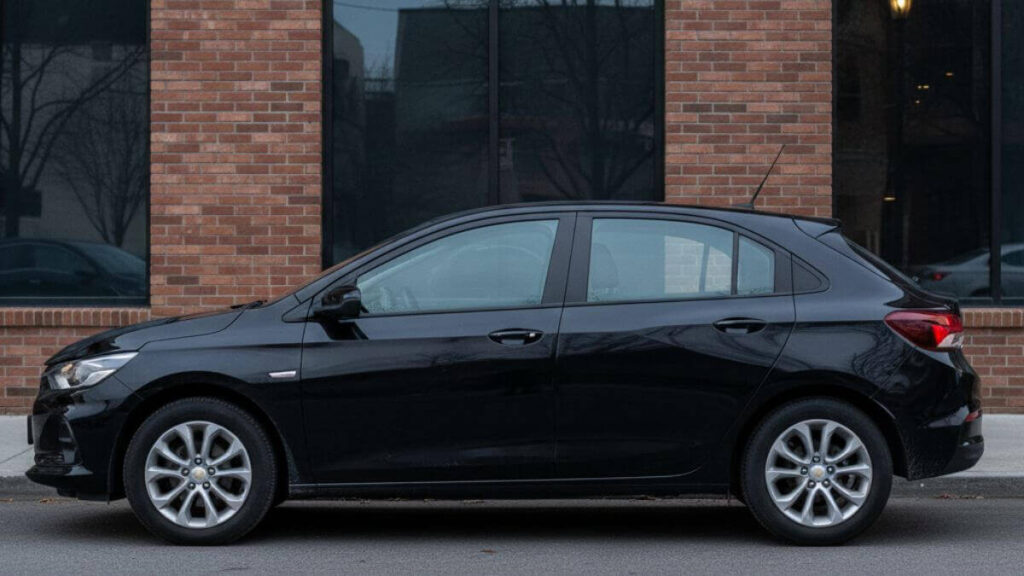 Vista lateral completa de um Chevrolet Onix preto estacionado em frente a um edifício moderno de tijolos, mostrando todo o design do carro, desde a frente até a traseira, em alta resolução.