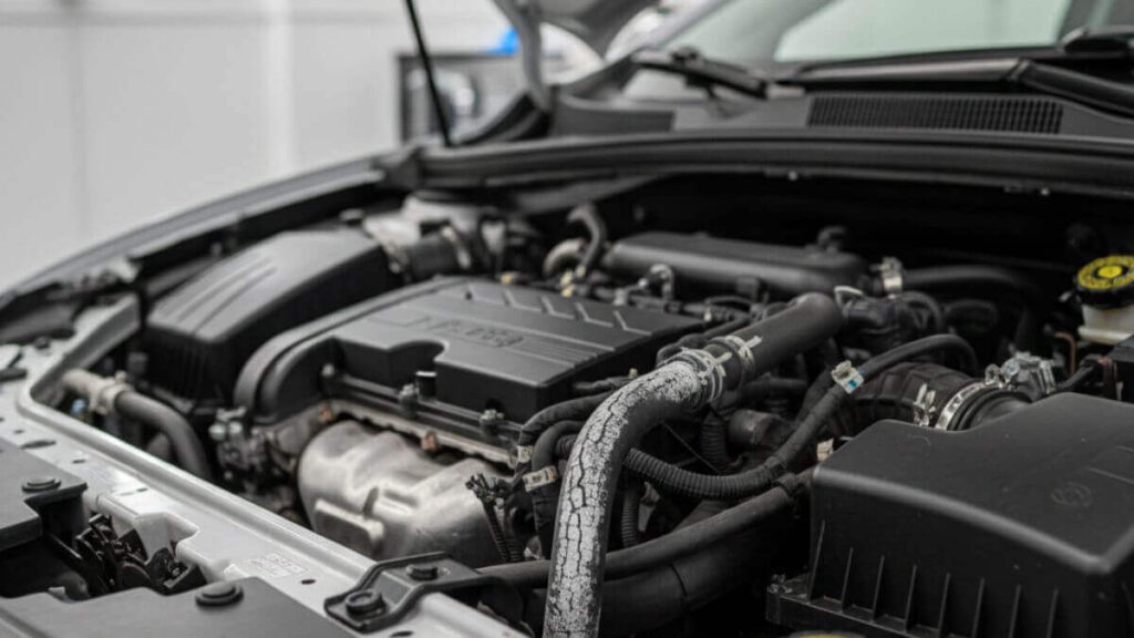 Close-up detalhado do compartimento do motor de um Chevrolet Onix com o capô aberto, focando em uma mangueira de borracha visivelmente ressecada e com rachaduras, indicando potencial entrada falsa de ar.