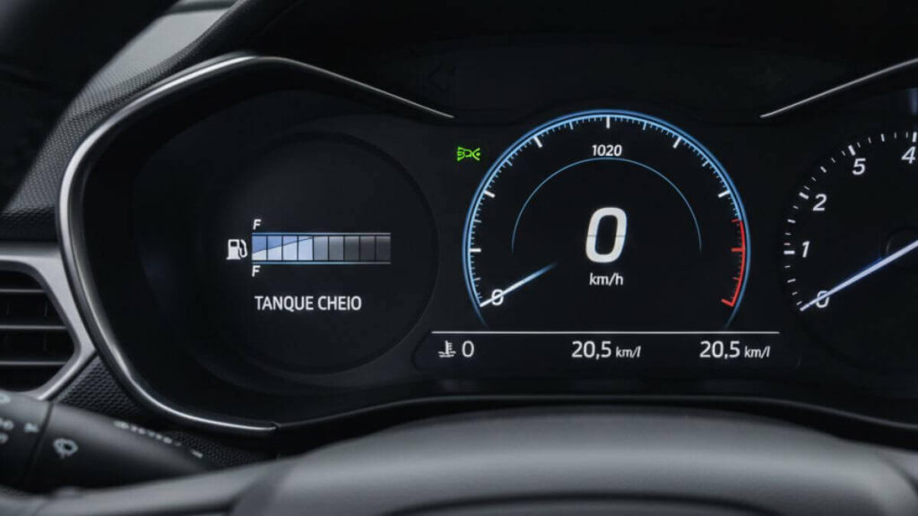 Close do painel digital de um Chevrolet Onix moderno, mostrando o marcador de combustível no nível máximo (tanque cheio) e computador de bordo indicando baixo consumo de combustível. Ao centro, o logotipo da Chevrolet no volante.