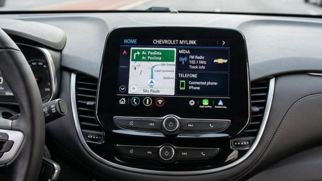 Multimídia original MyLink Foto profissional do interior de um carro Chevrolet modern (como Tracker ou Onix), capturada do ponto de vista do motorista. Mostra o volante com o logotipo Chevrolet à esquerda e a central multimídia MyLink ao centro da consola. A tela exibe um mapa de GPS (São Paulo, Rua da Consolação), informações de rádio e status do telefone. Ao fundo, uma avenida movimentada e prédios da cidade.