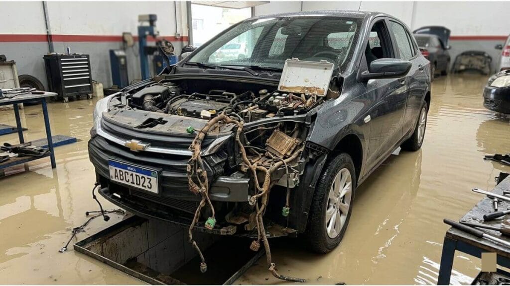 Um Chevrolet Onix cinza com o para-choque dianteiro removido está estacionado em uma oficina mecânica com o chão coberto por água barrenta. Fios elétricos e conectores estão soltos e pendurados na parte frontal do motor e do chassi. Uma caixa branca de fusíveis está aberta e visivelmente danificada.