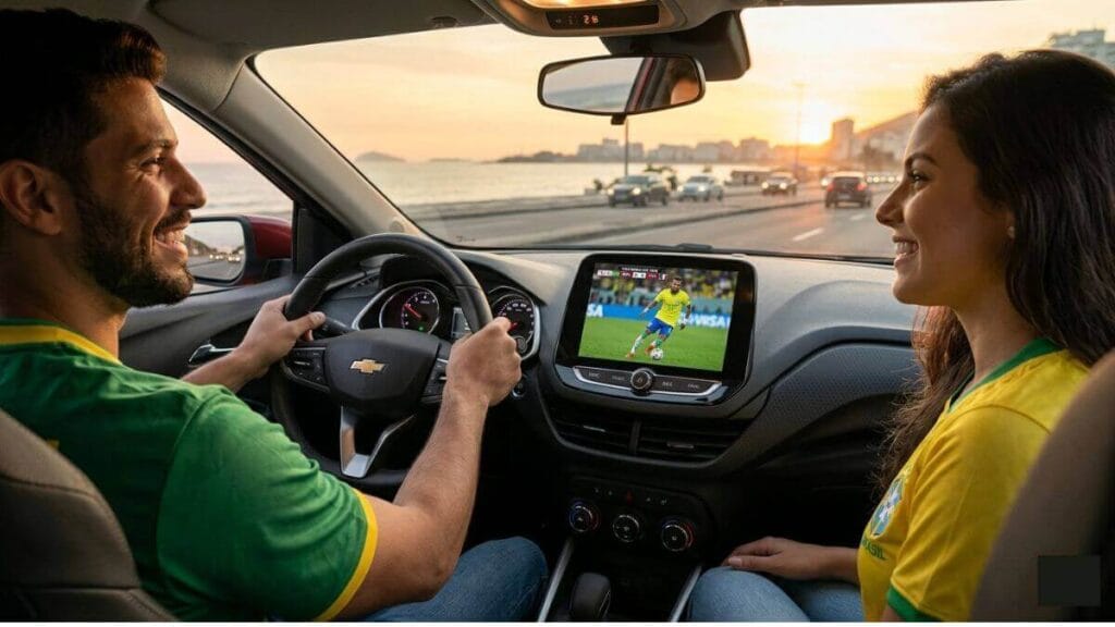 Casal a bordo de um Chevrolet Onix com a central multimídia conectada exibindo um jogo de futebol da Copa do Mundo ao vivo; o motorista sorri mantendo a atenção na estrada enquanto a passageira assiste à transmissão.