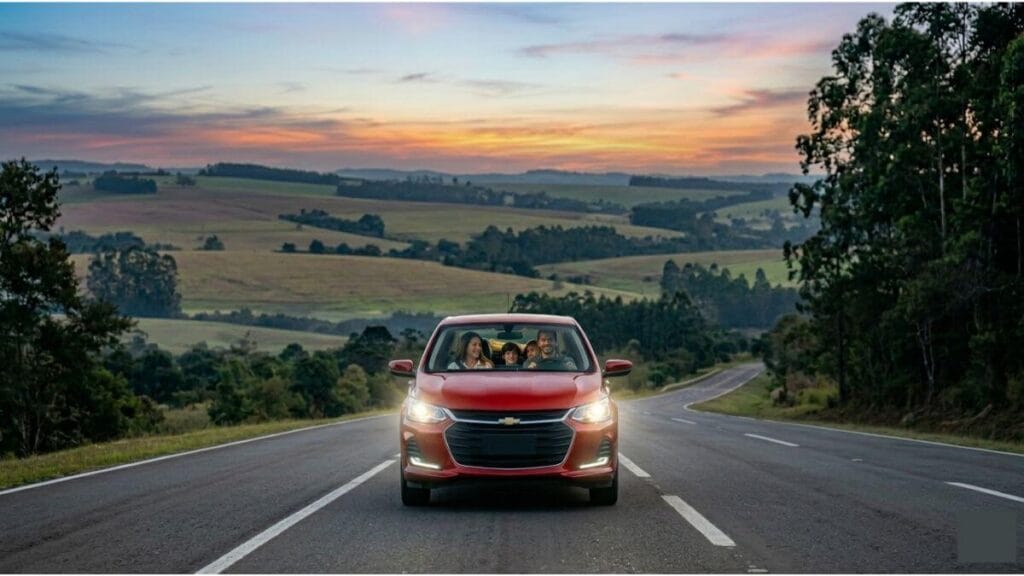 Família sorridente dentro de um Chevrolet Onix vermelho na estrada, com os faróis acesos, pronta para viajar em clima de Copa do Mundo.