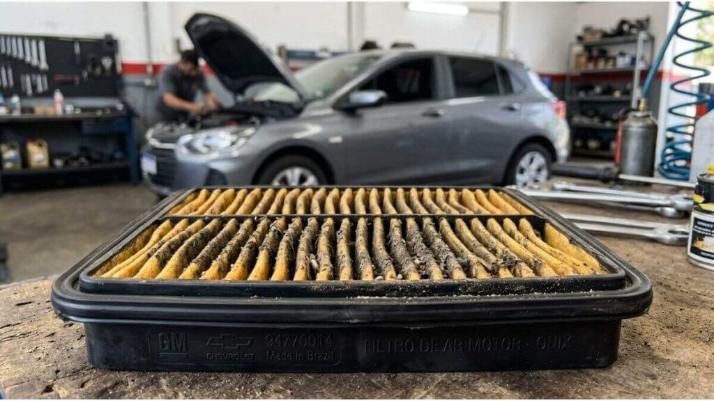 Filtro de ar do motor de um Chevrolet Onix acumulado com sujeira e poeira em destaque sobre uma bancada, com um Onix cinza ao fundo em uma oficina.